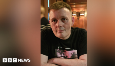 A man is sitting in a restaurant. He's wearing a black top with a design showing palm trees against a sunset. He has brown hair. A woman is passing in the background. Some light fixtures are on the wall.