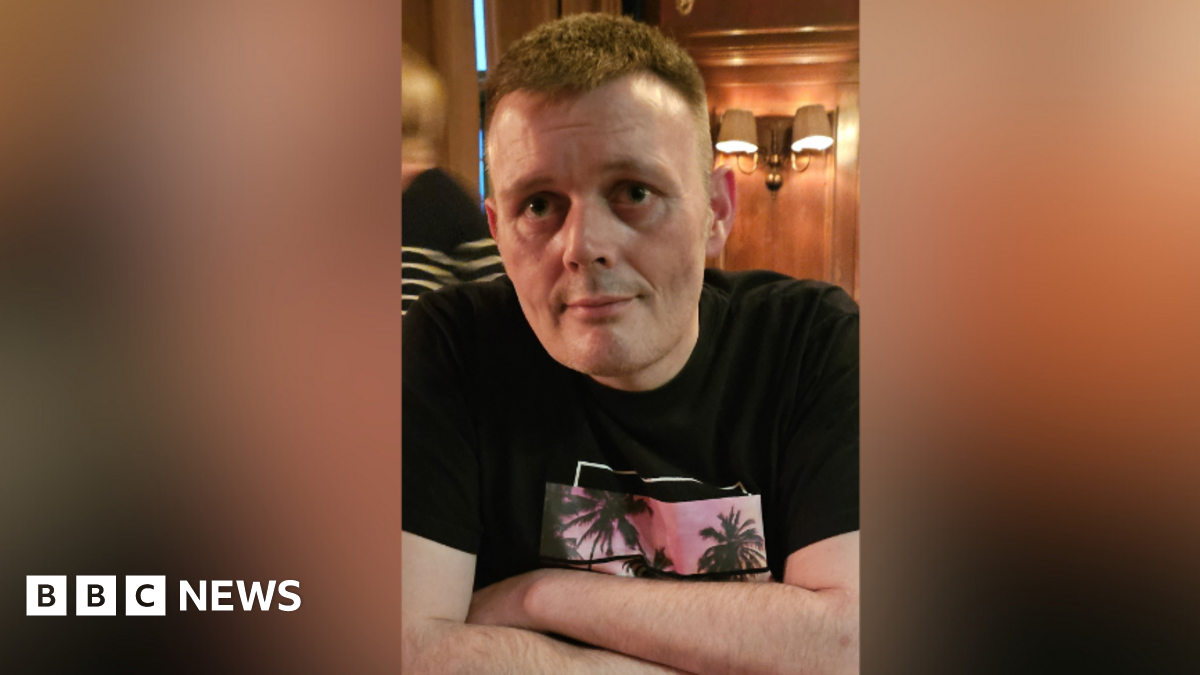 A man is sitting in a restaurant. He's wearing a black top with a design showing palm trees against a sunset. He has brown hair. A woman is passing in the background. Some light fixtures are on the wall.