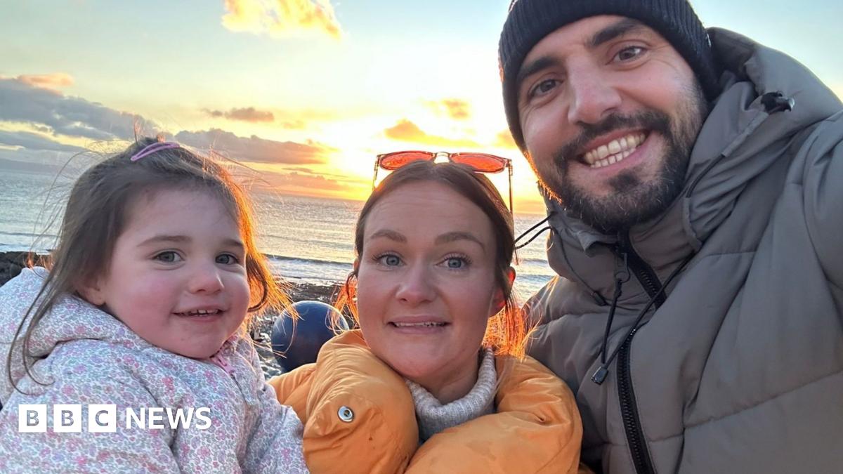A woman and her husband pose with their young daughter, all wearing coats, at the seaside.