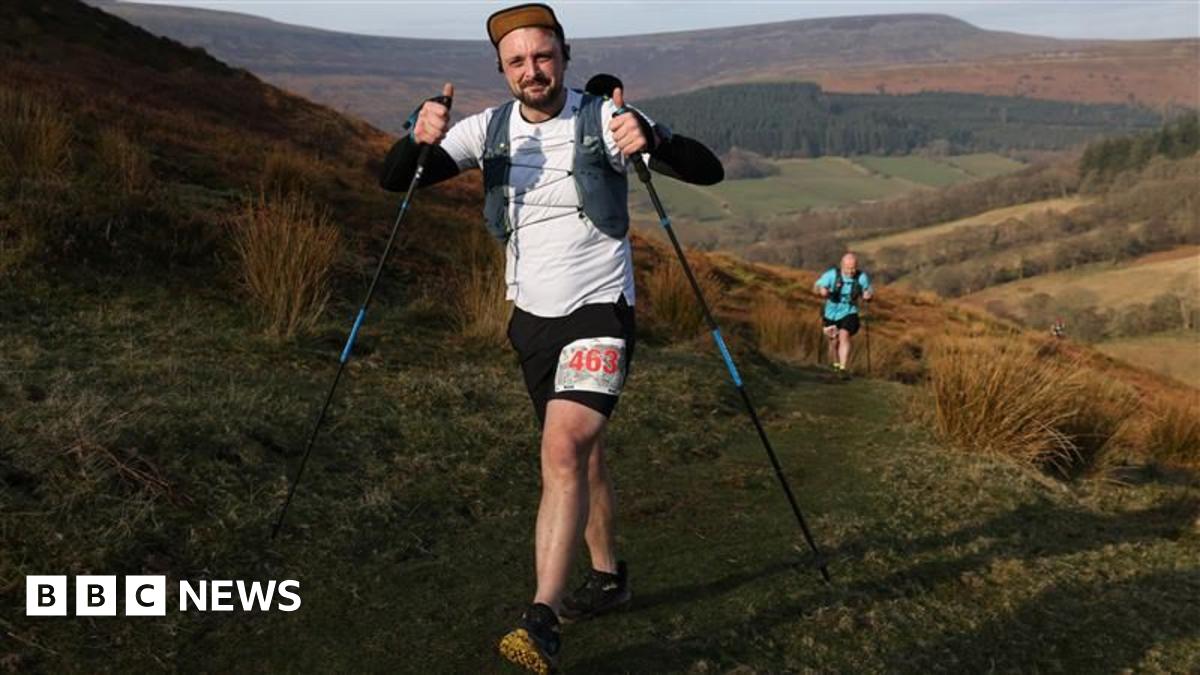 A man in a white top running up a steep hill, he is holding two poles and wearing a cap.