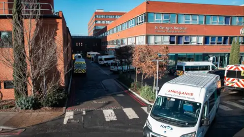 Getty Images Several ambulances are parked outside St George’s Hospital in Tooting, South London. The large red-brick building has multiple windows and a sign reading ‘Grosvenor Wing Main Entrance’ above the entrance.
