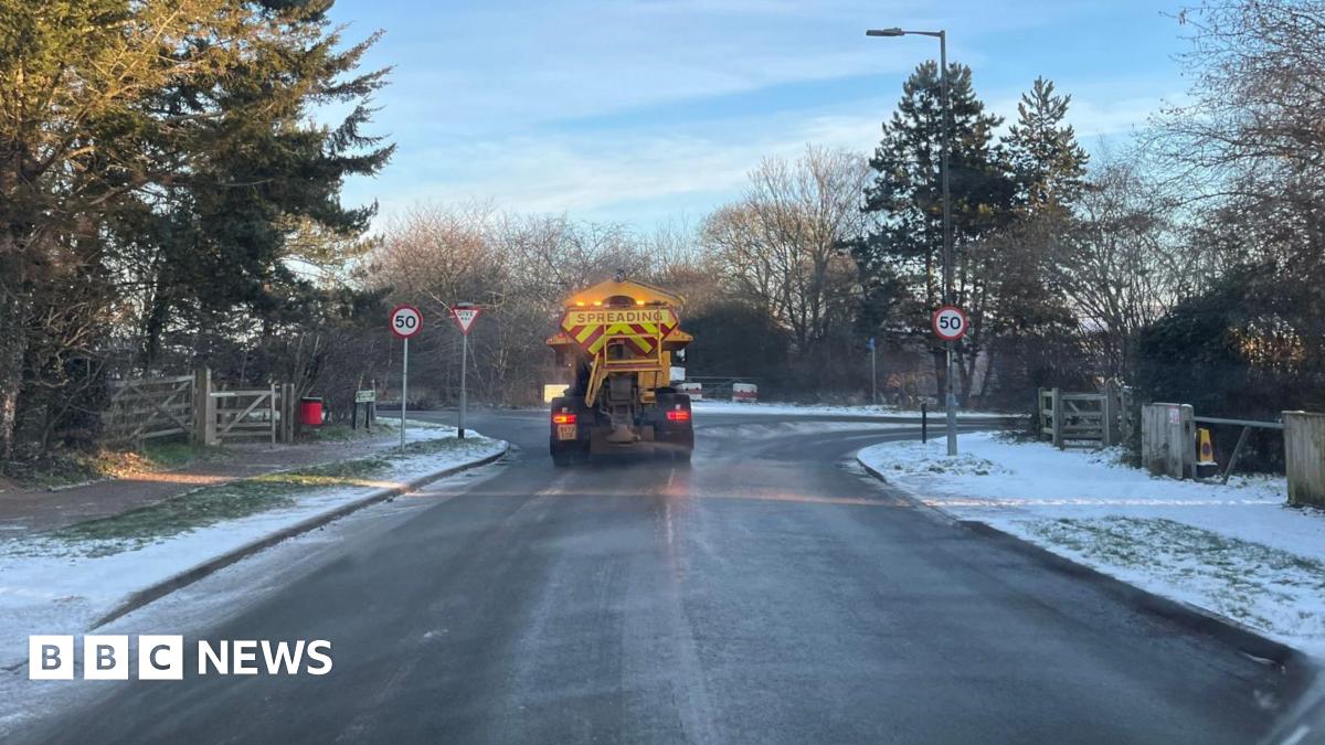 The back of a yellow gritter lorry, with a yellow and red striped sign that says "Spreading". It is at a T-junction, with the verges mostly covered in snow.