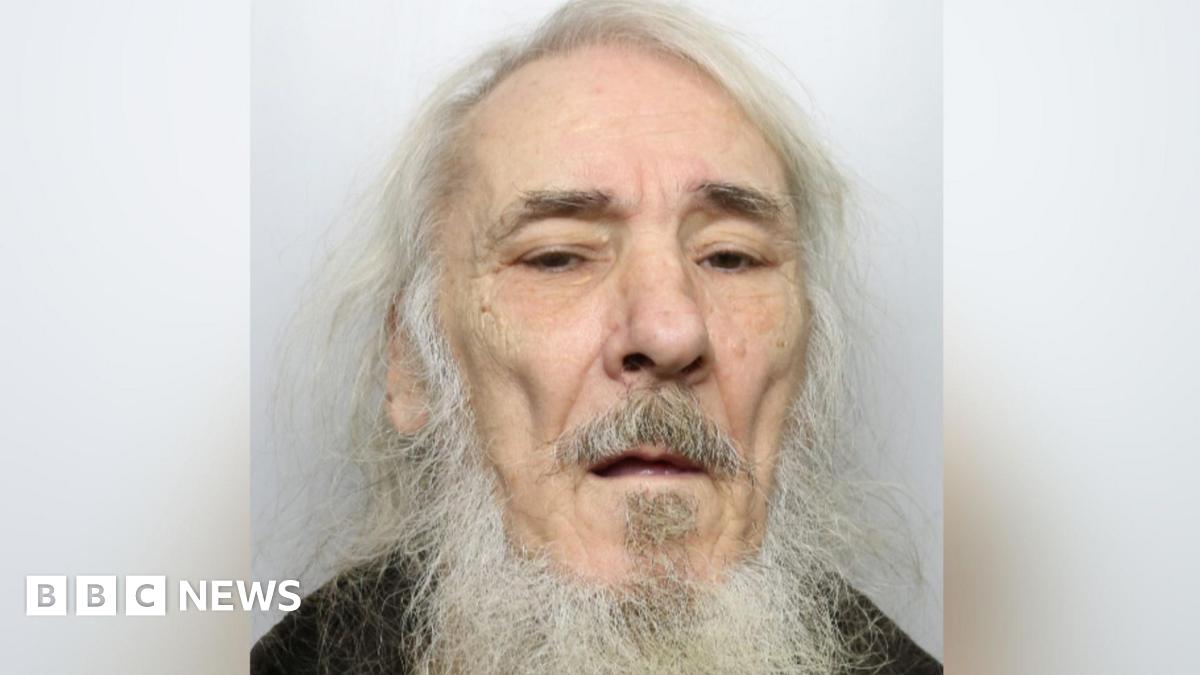 An elderly man with long white hair and beard. He is standing against a grey background wearing a black top.