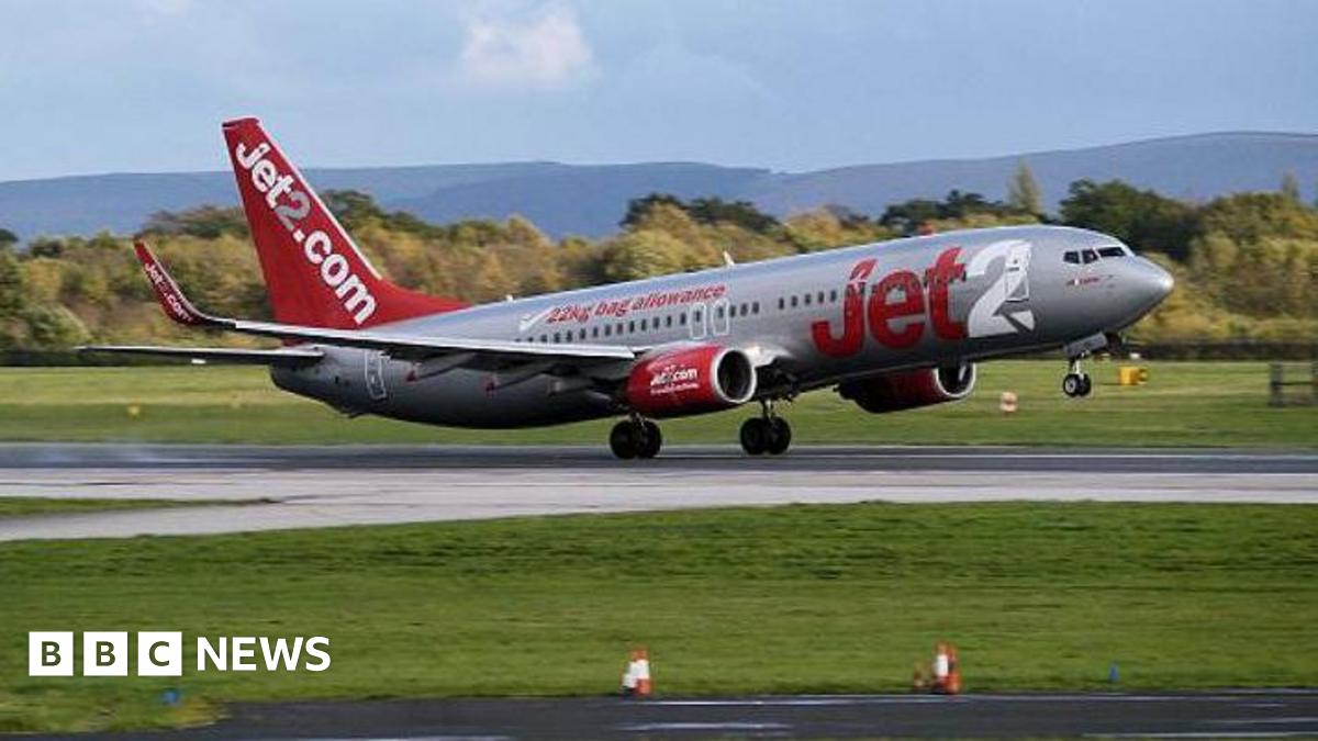 Jet2 aircraft takes off from a runway at Manchester Airport on a clear day. Hills of the Derbyshire Pennines in the background.
