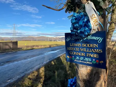 A blue sign with the words In Loving Memory and the names Lewis Soden, Reece Williams and Connor Page attached to a tree by the side of a rural road