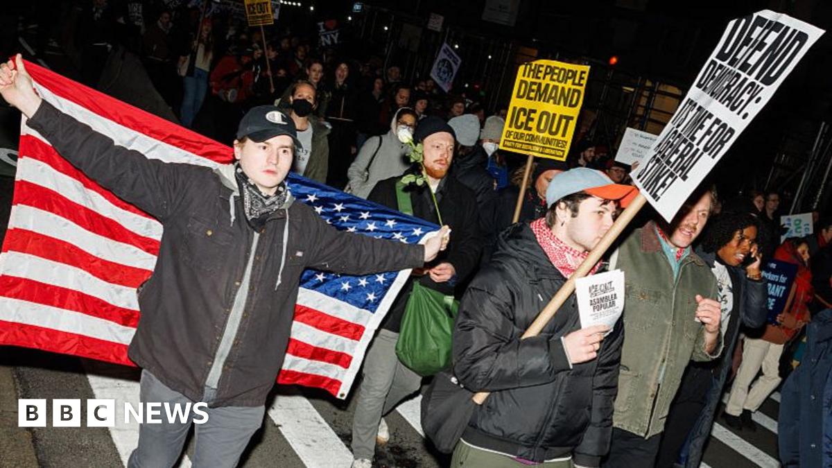 Three people are standing together in support for Renee Nicole Good. One man is flying a Mexican flag, the woman in the middle is visibly crying, and a man on the right is holding his fist up.