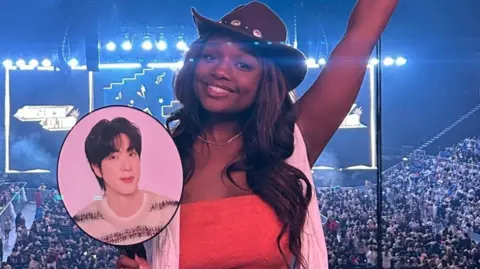 Lauryn Adene Lauryn is wearing a brown cowboy hat, she has brown long wavy hair. She is wearing a red top with a white scarf. She is stood with her back to the stage and concert full of people, holding a face card with BTS member Jin on it. 