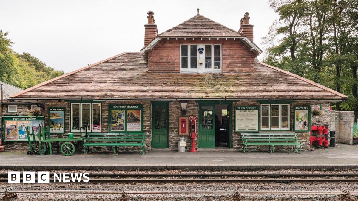 Woody Bay Station viewed from across the railway tracks. It is in the style of a Swiss chalet and has two chimneys, a slanted roof and green doors. There are green benches outside it and old-style luggage carrying trolleys. There is also a post box built in to the front wall. There are trees beyond.