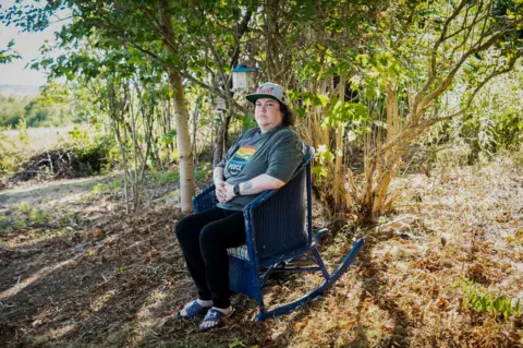 Jillian Lucas sits in a blue wicker rocking chair in her back yard, under a tree bathed in sun. 
