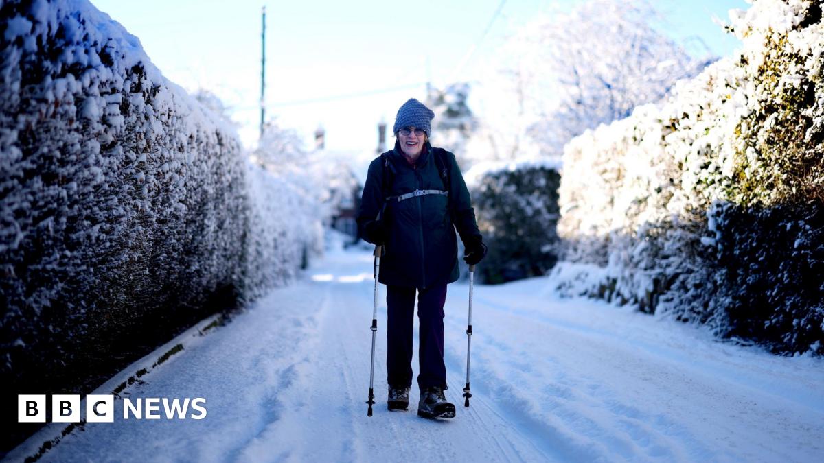 A woman walks through a snowy lane wearing a wooly hat, using poles.