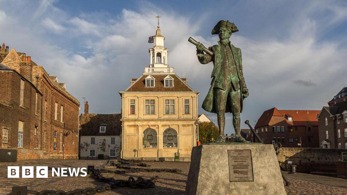 A statue of Captain Vancouver stands on a plinth. The bronze statue is wearing a tricorn and clutching a scroll. Behind him is the Custom House, a 17th century three storey building, topped by a white tower.