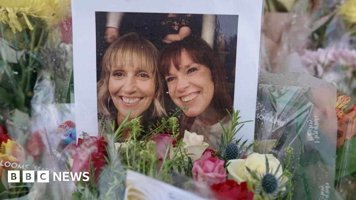 A photograph showing two women - one with blonde hair and one with brown hair - is propped up against bouquets of brightly-coloured flowers.