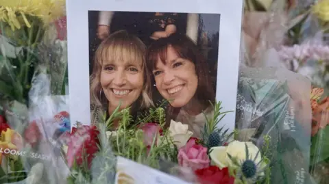 BBC A photograph showing two women - one with blonde hair and one with brown hair - is propped up against bouquets of brightly-coloured flowers.