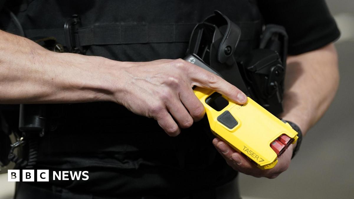 Stock image of an officer demonstrating a new Taser. It is a close up shot of a man's hands holding a yellow taser, with the name Taser 7 on it and there are two red cartridges visible.