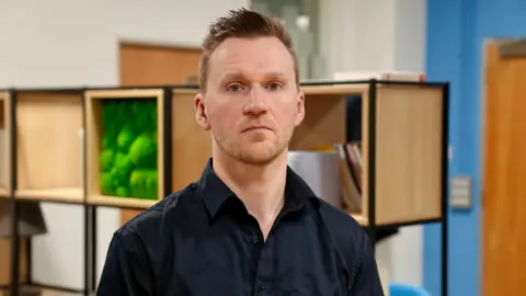 Joe looking at the camera with a straight expression. He has short light brown hair, brown eyes and stubble. He is wearing a navy blue shirt and standing in front of a display case made of large cubes which is blurred in the background. 