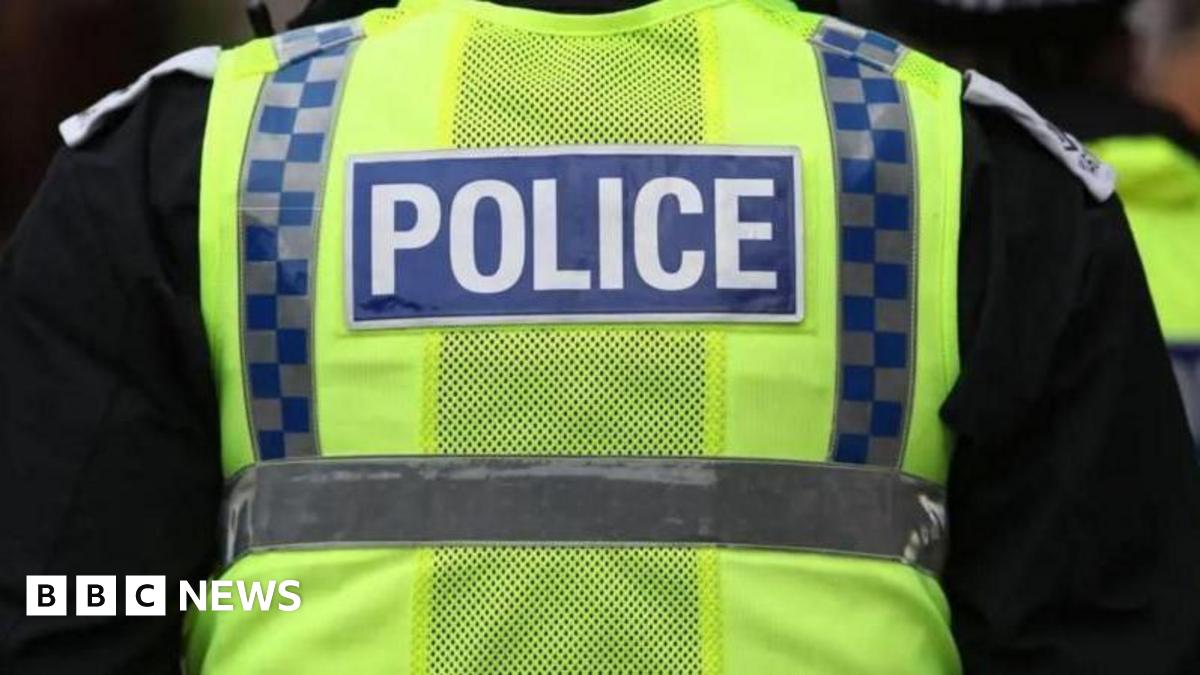 A stock image of the back of a police officer wearing a yellow hi-vis vest with blue and grey checks running down the sides. Police is written in the middle of the vest in white letters in a blue box. The officer is also wearing a black long-sleeved top underneath.