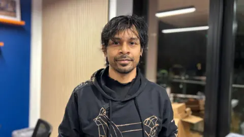 BBC Man wearing a dark grey hoodie looks at the camera. He has black hair, slight brushed forward fringe and a short beard growth. He is in what looks like an office, with a window looking out to a balcony.