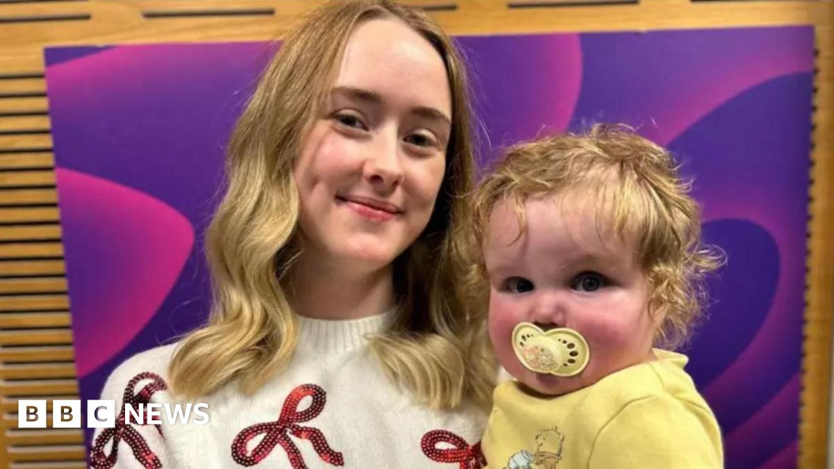 A woman with long blonde hair and a white jumper with red bows holding a baby with blonde curly hair, a yellow dummy and a yellow top in front of a wood-panelled background with a purple sign on it