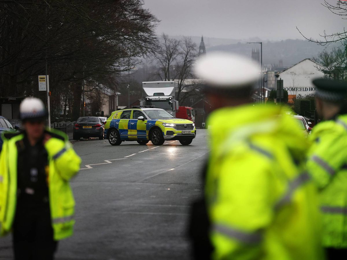 Emergency response personnel in high-visibility jackets are gathered on a street, with an ambulance car in the foreground. The scene is set on a cloudy day with distant mountains in the background.