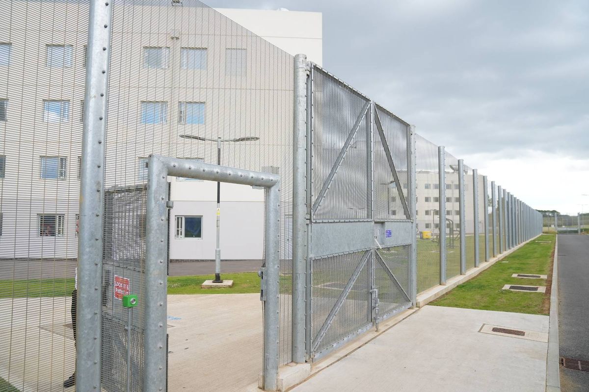An exterior view of a facility featuring a tall, white building with multiple windows, a paved entrance with an open metal gate, and a perimeter fence. The surroundings include a grassy area and a clear sky.