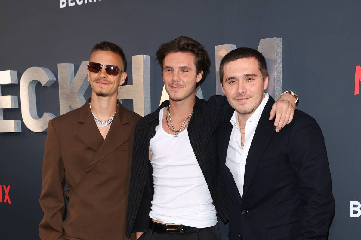 Three individuals, dressed in formal attire, are standing together on a carpeted surface. They appear to be at a premiere event, as indicated by the wall in the background that features a large letter "E" and the word "ECK" prominently displayed. The two individuals on the right are closely positioned, with one placing a hand on the shoulder of the other, suggesting a camaraderie or connection between them.