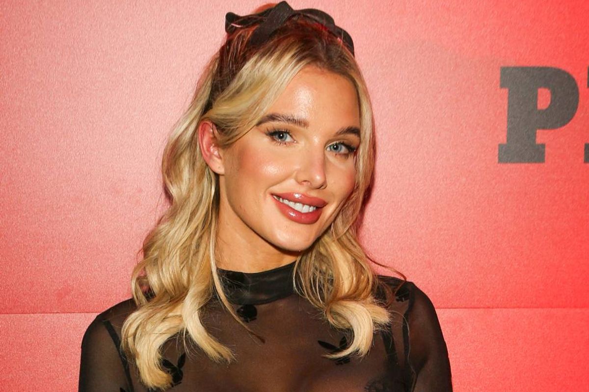 An individual with blonde hair is standing in front of a red wall, smiling and looking directly at the camera. The wall bears the letter "P" prominently displayed.