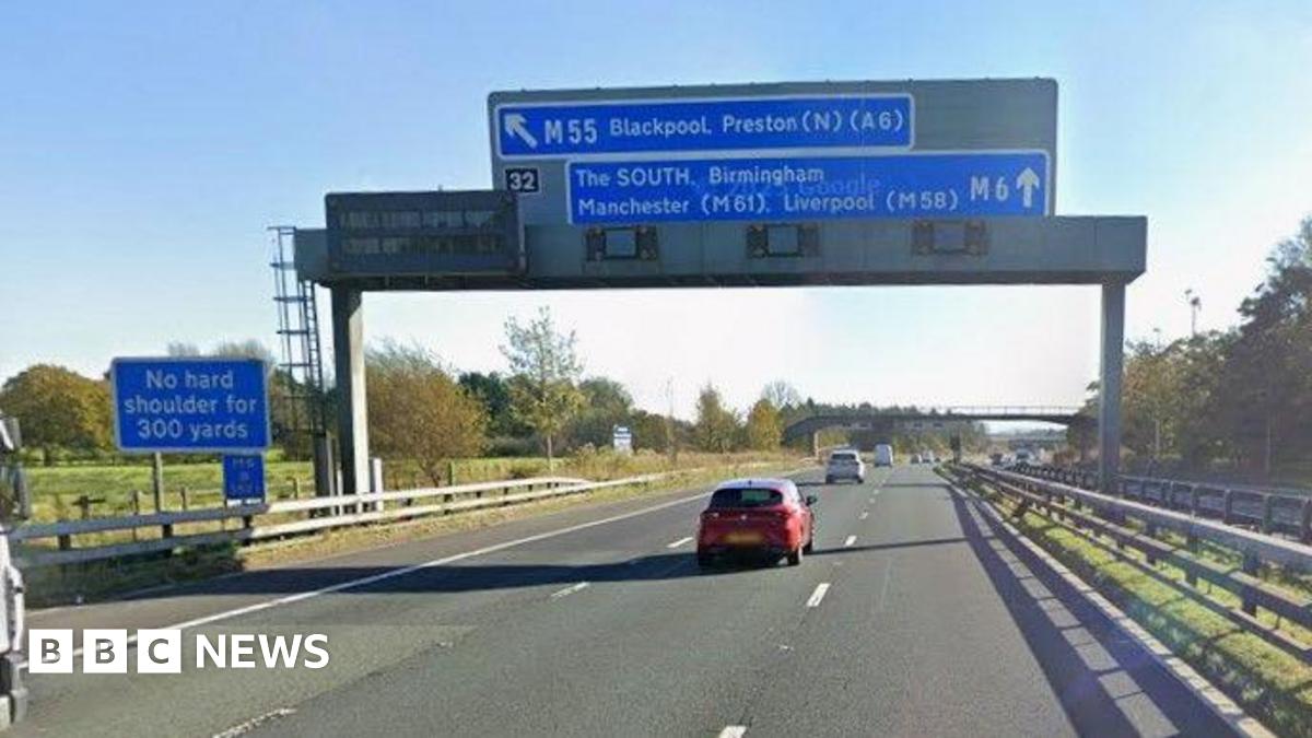 A blue sign on a board over a motorway indicating a slip road for the M55 towards Blackpool and Preston