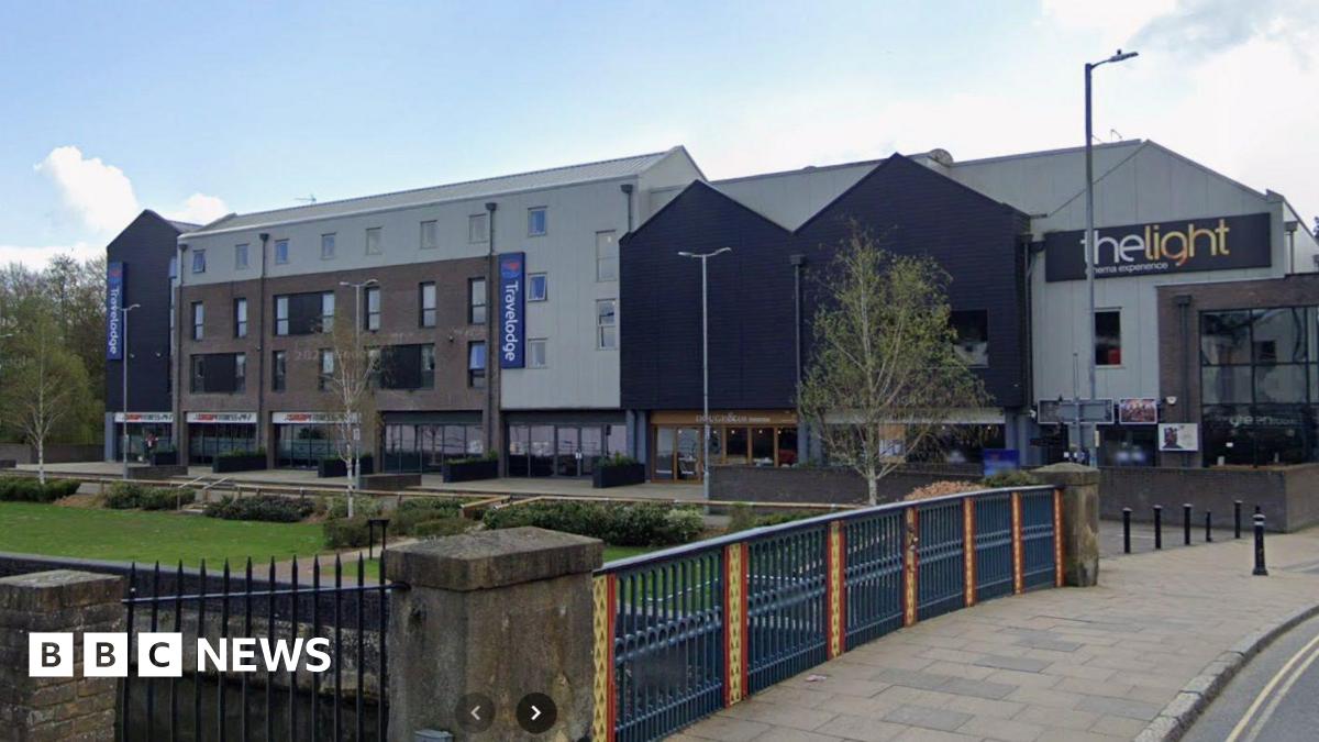 A number of connected buildings, including a Travelodge and The Light cinema, are set back from the grassy bank of a river. In the foreground, a bridge crosses the river.