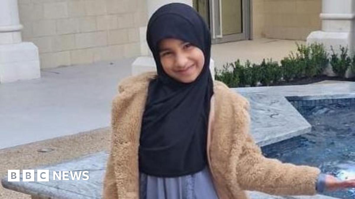 A young girl, wearing a brown furry jacket and a headscarf smiles at the camera as she stands next to a water fountain in the shape of a star.