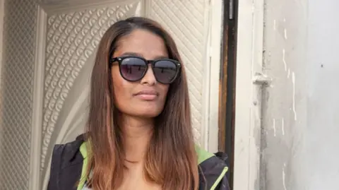 Sam Tarling/Getty Images Shamima Begum leaning against a door frame. She has long dyed brown hair and is wearing sunglasses with a light grey top underneath a sweatshirt jacket