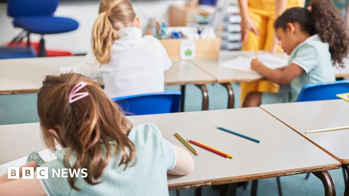 Schoolchildren sit at desks in a classroom, writing on paper with pencils. A teacher stands nearby, while coloured pencils lie on the tables in the foreground.