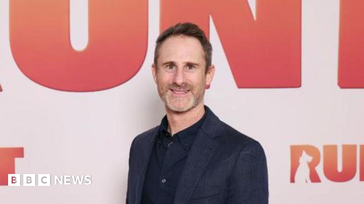 Millie Bobby Brown and David Harbour in character as Eleven and Jim Hopper. Eleven wears red shorts over grey leggings and a grey jumper. Jim Hopper wears a black leather jacket, a black beanie hat and dark trousers.They are speaking to director Frank Darabont on the set of Stranger Things. There is a vintage advertisement posted in the background with debrief on the ground.