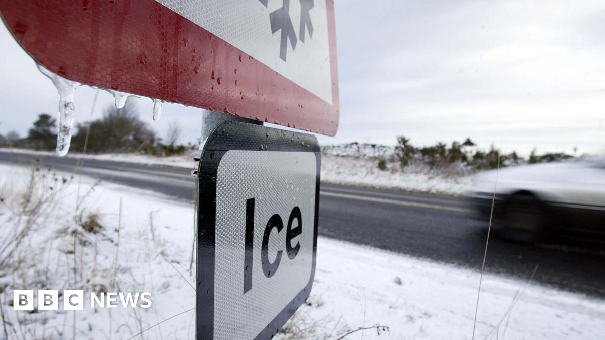 A stock image of a road sign warning of ice in the foreground, with a speeding car on a snowy road behind it.