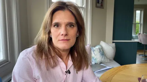 BBC Ellen Roome, sat in her living room, looking at the camera. She has brown mid-length hair and a pink and white shirt on