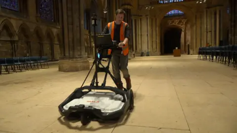 BBC A man pushes a radar device on wheels down the nave of a cathedral. The device is shaped like a lawnmower with a black frame and handles, wheels and a white cover in the centre. The man is wearing a black shirt, grey trousers and an orange hi-vis vest. The cathedral has sand-coloured paving stones and stone walls and pillars.