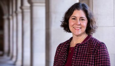 A portrait of Beverly Wendland who is standing in front of long row of columns wearing a maroon shirt and patterned jacket.