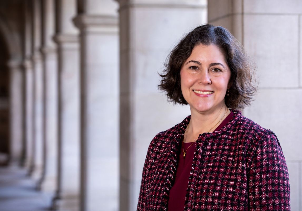 A portrait of Beverly Wendland who is standing in front of long row of columns wearing a maroon shirt and patterned jacket.