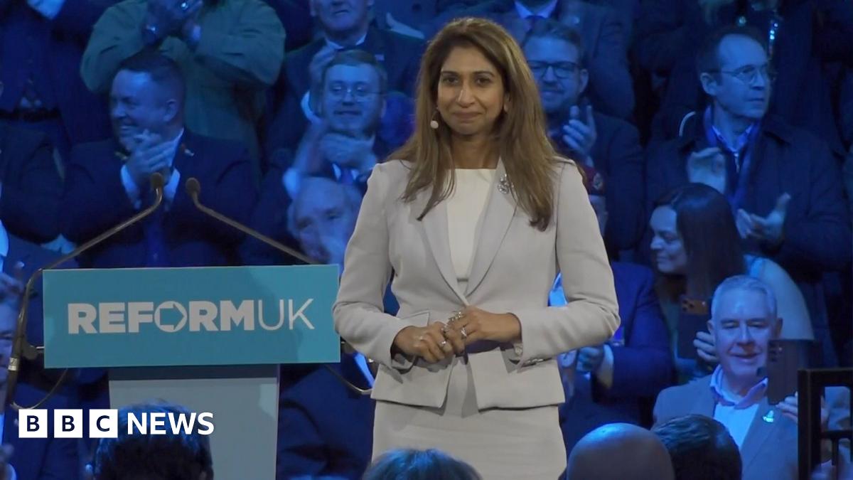 Suella Braverman in a grey and white suit beside a speaking lectern with Reform UK to her left. Audience is visible behind her lit by blue-purple lighting.