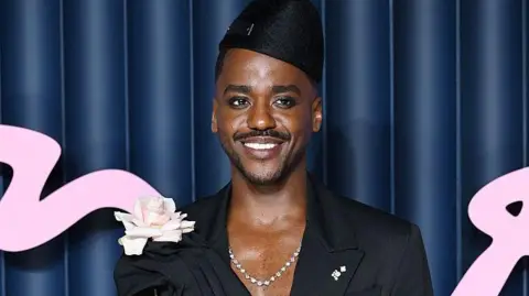 Getty Images Gatwa wearing a stylish black draped outfit with a deep neckline, accessorized with a silver necklace and a black headpiece, holding a single long-stemmed pale pink rose pinned to the left side of the outfit. The background features dark blue vertical panels with large pink decorative text