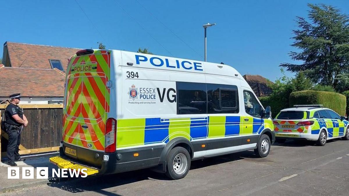 A white police car and van parked on a residential road, which has a large tree, a bush and a house on it. A police officer guards the entrance to a property.