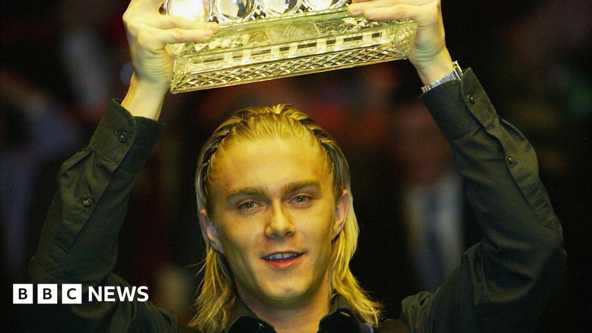 The snooker player Paul Hunter, lifting a trophy aloft out of shot. His blond hair is braided and he is wearing a black shirt.