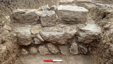 Mike Luke A Roman wall, with a red and white stick in front of it. There is soil all around it and tools to the right. 