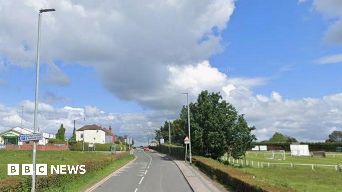 A rural road with fields on both sides. Lamp-posts and trees can also be seen.