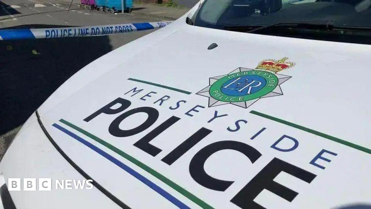 The white bonnet of a Merseyside Police patrol car, bearing the name of the force in black and blue lettering. Police tape can be seen across a road behind the vehicle.