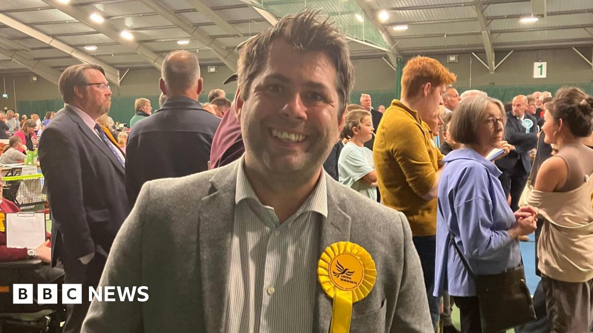 Leigh Frost wearing a grey blazer with a white striped shirt. He also has a yellow rosette on his left side with the Liberal Democrat bird logo on it. He has short brown hair and is stood in front of a crowd of people at the count.