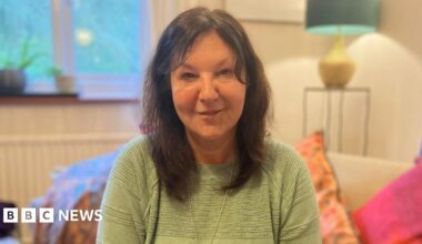 A dark-haired woman in a light green top. She is sitting on a sofa with several colourful pillows.
