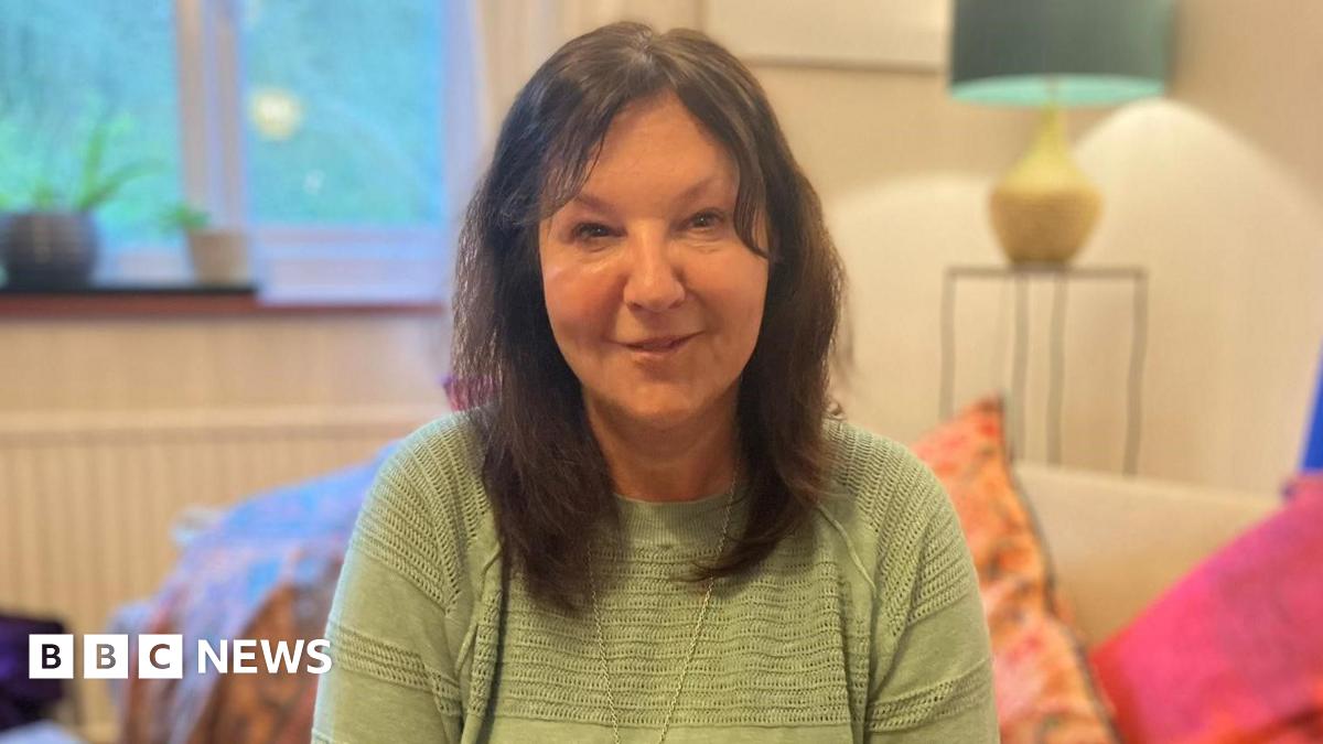 A dark-haired woman in a light green top. She is sitting on a sofa with several colourful pillows.