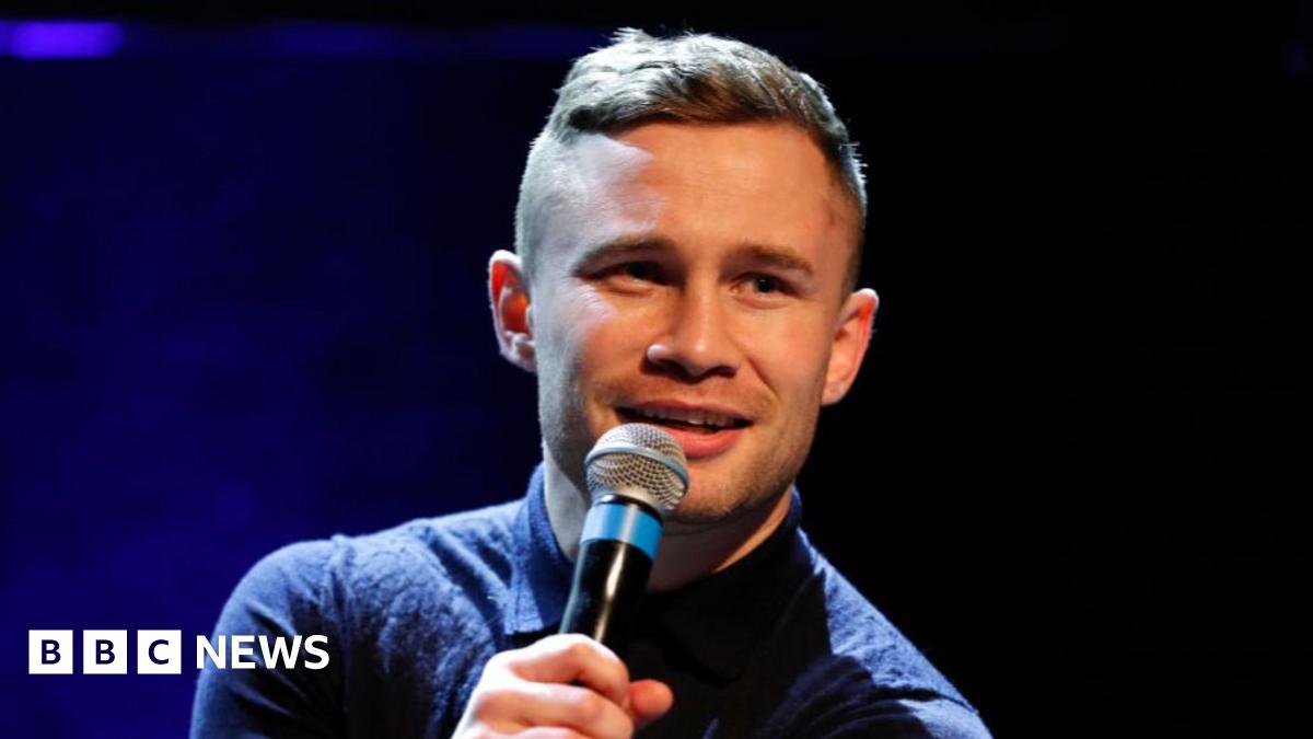 Carl Frampton has short brown hair and is talking into a microphone. He is wearing a navy polo shirt. The background is a black and blue stage.