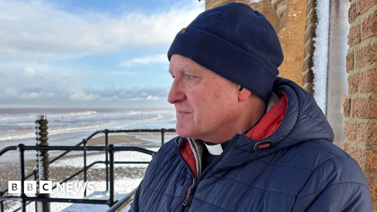 A reverend, wearing a black shirt with white dog collar under a navy jacket, looks out to sea. He has a sombre, concerned expression.