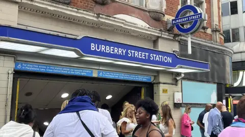 People walking past the entrance of a London Underground station with a blue sign reading “Burberry Street Station” above the ticket hall entrance.